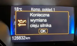 Wyświetlacz samochodowy z komunikatem "Konieczna wymiana oleju silnika" i ikoną klucza.