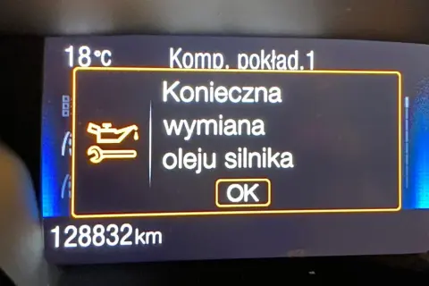 Wyświetlacz samochodowy z komunikatem "Konieczna wymiana oleju silnika" i ikoną klucza.