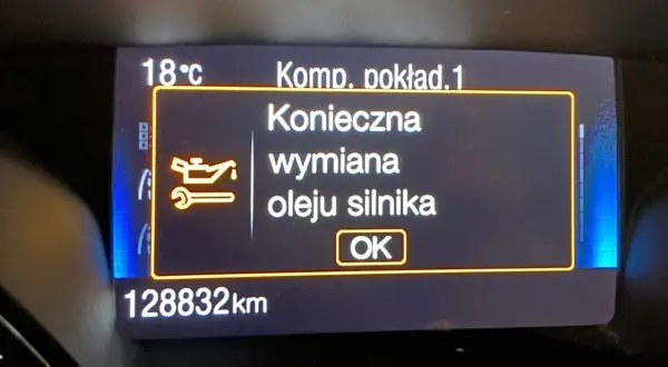 Wyświetlacz samochodowy z komunikatem "Konieczna wymiana oleju silnika" i ikoną klucza.